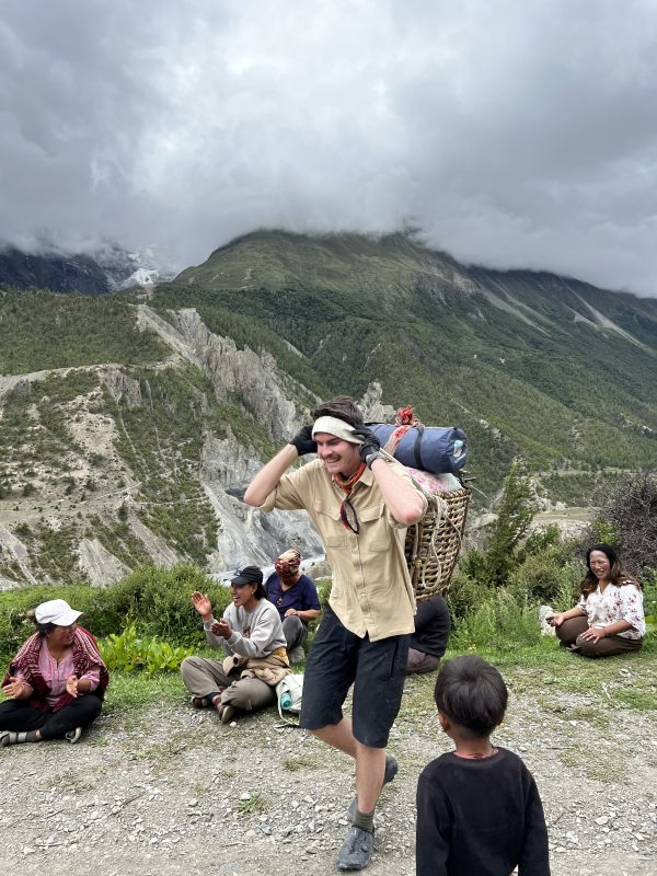 A man carries camping supplies on the back of his shoulders along a mountain trail, with a valley and more mountains in the background. Women and children sit along the trail, smiling and clapping.