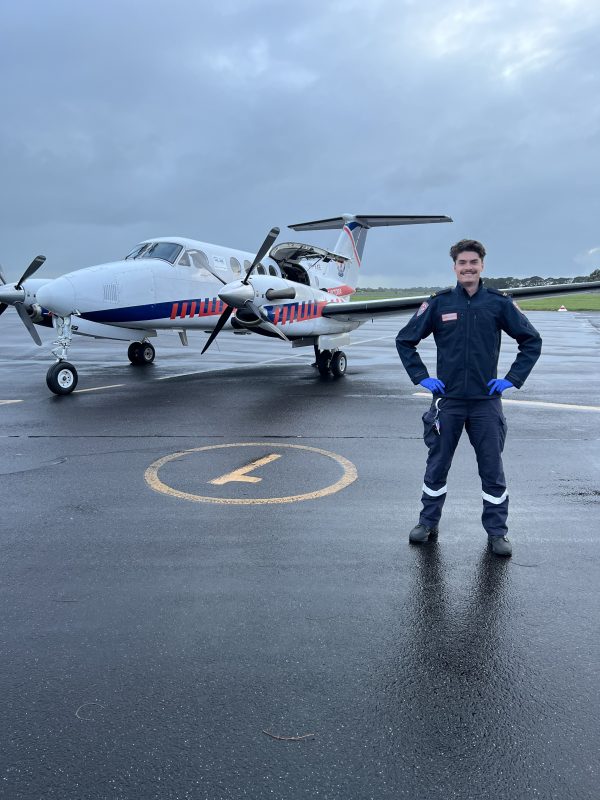 A man in Ambulance Victoria on-road uniform smiles for the camera in front of an ambulance plane.