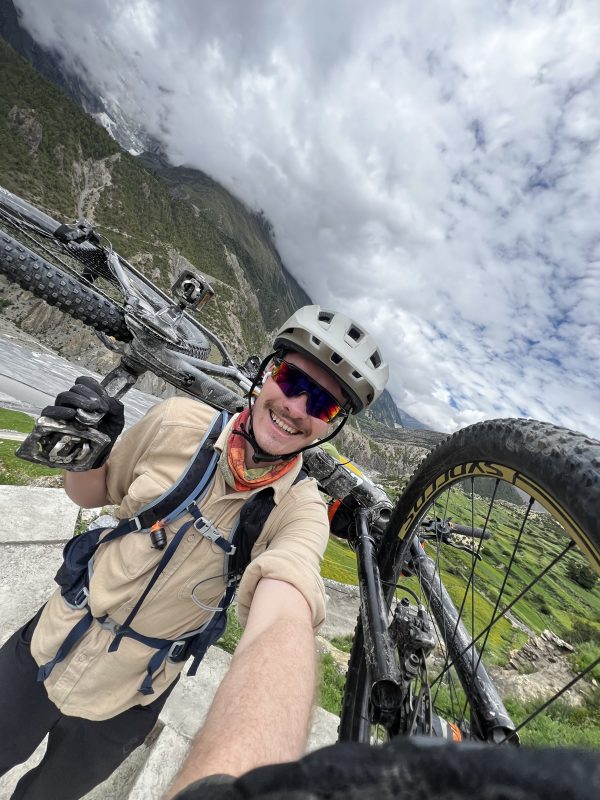 A man takes a selfie with his mountain bike on his shoulders. Mountains are in the background.