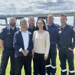 A man and a woman stand in front of four uniformed emergency service workers. The group smile for the camera. They stand outside on grass and a big road bridge is in the background.