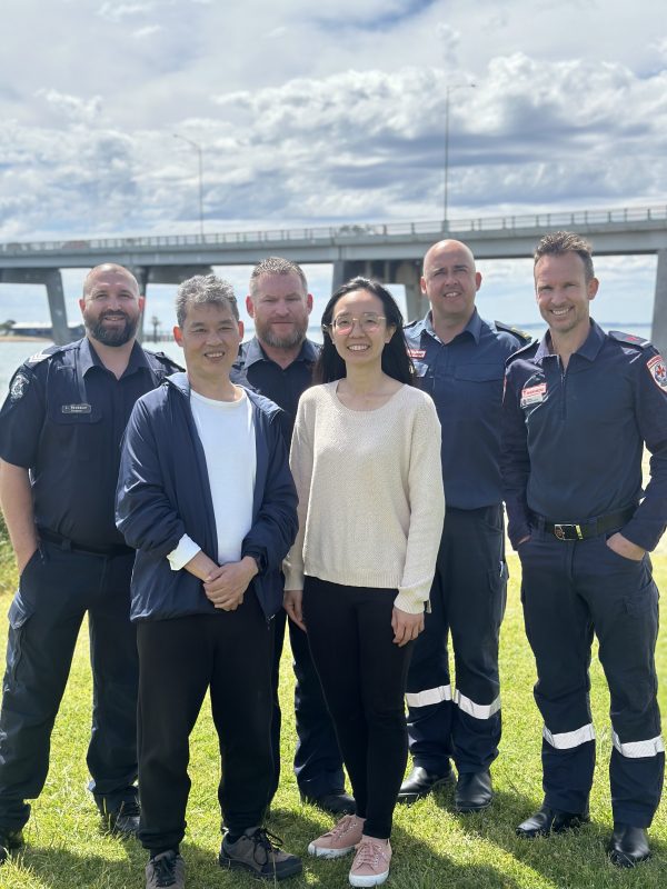 A man and a woman stand in front of four uniformed emergency service workers. The group smile for the camera. They stand outside on grass and a big road bridge is in the background.