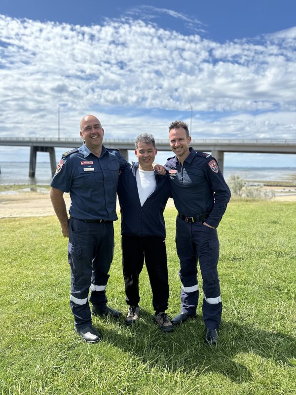 Two paramedics smile with their arms around a man in casual clothes. They stand on grass with a small beach, water and a road bridge in the background.