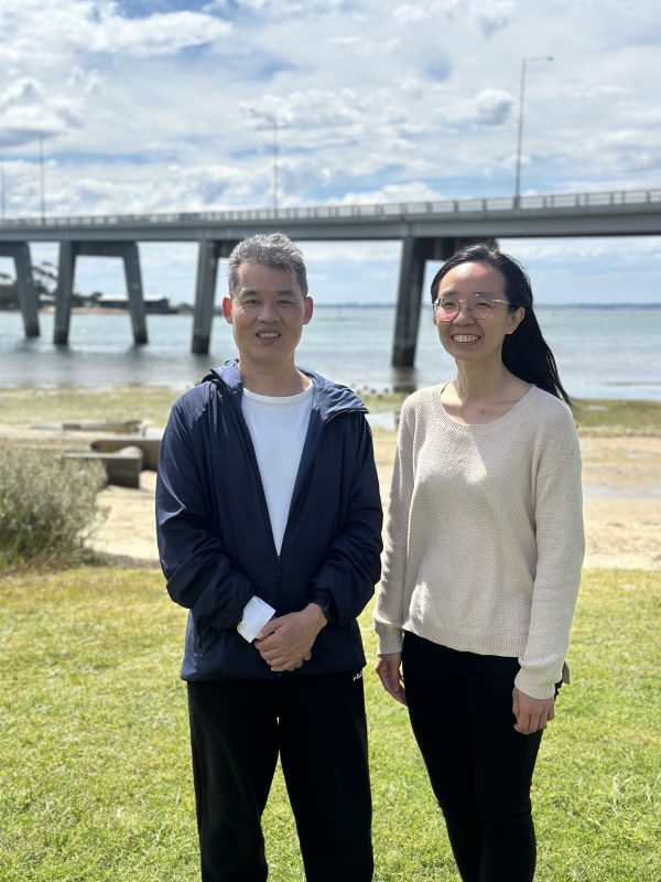 A man and a woman stand side-by-side and smile for the camera. They are outside and a small beach, water and a road bridge are behind them.