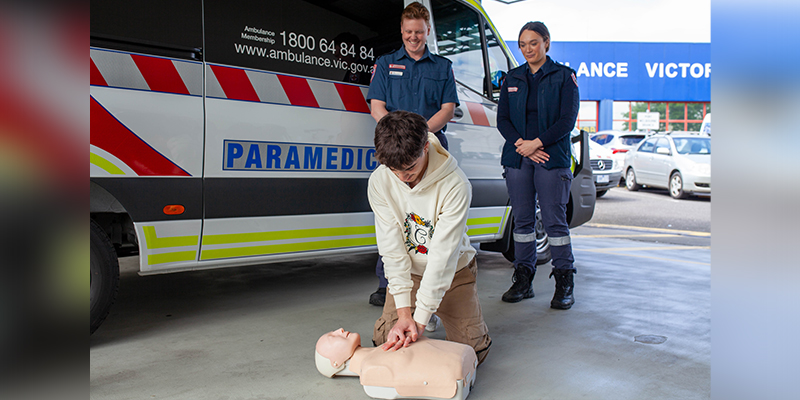 A teenage boy is showing how to perform cardiopulmonary resuscitation on a manikin beside an ambulance while two paramedics look on.