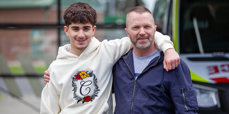 A teenage boy is standing beside an older man and has his left arm over his shoulders.