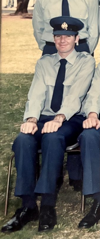 A young man sitting on a chair smiles for the camera in RAAF formal wear - a peaked cap, collared shirt, tie, navy pants and dress shoes. The image is cropped to focus on the man, but would have been a posed group photo.