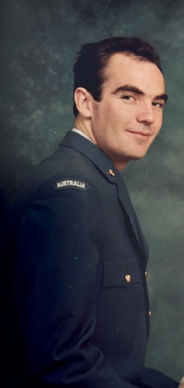 A young man looks over his shoulder and smiles at the camera. It's a colour portrait shot. He wears a navy blue jacket which says 'Australia' on the shoulder.