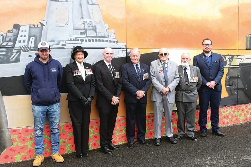 A group of men and women stand side by side in front of a wall with a mural painted on it. Poppies are painted along the bottom of the wall. Five of the seven people have medals pinned to their jackets.