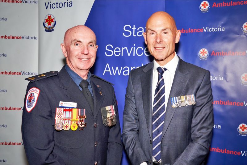 Two men smile for the camera with medals pinned to their jackets. The man on the left wears Ambulance Victoria uniform. The man on the right is in a suit. Behind them, a banner indicates they are at the Ambulance Victoria Staff Service Awards.