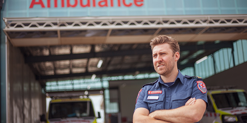 A man standing with his arms folded outside a garage with two ambulance vehicles parked in it.