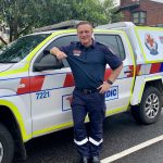 A man paramedic smiles for the camera, leaning against an Ambulance Victoria car.