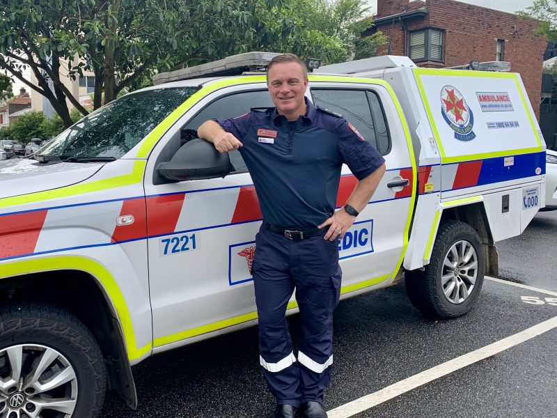 A man paramedic smiles for the camera, leaning against an Ambulance Victoria car.