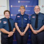 Two women and a man in Ambulance Victoria formal uniform smile for the camera, in front of Ambulance Victoria banners.