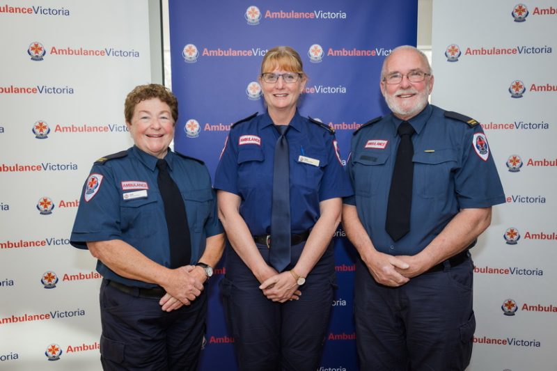 Two women and a man in Ambulance Victoria formal uniform smile for the camera, in front of Ambulance Victoria banners.