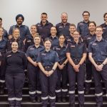 A group of 20 people in Ambulance Victoria uniform smile for the camera. They stand in four rows on stairs.