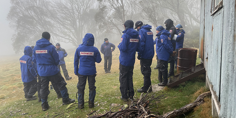 A group of people wearing blue paramedic jackets are standing in a circle outside a hut in the forest.