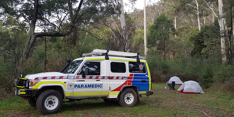 A vehicle is parked in the middle of a forest. There are two tents set up behind the vehicle.