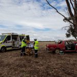 Ambulance Victoria First Responders wheel a patient on a stretcher towards an ambulance. A car appears to have crashed into a tree in a rural area.