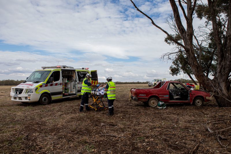 Ambulance Victoria First Responders wheel a patient on a stretcher towards an ambulance. A car appears to have crashed into a tree in a rural area.