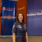 A woman in Ambulance Victoria formal uniform smiles for the camera in front of Ambulance Victoria banners and balloons.