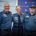 Three people in Ambulance Victoria paramedic uniform smile for the camera, in front of Ambulance Victoria banners.