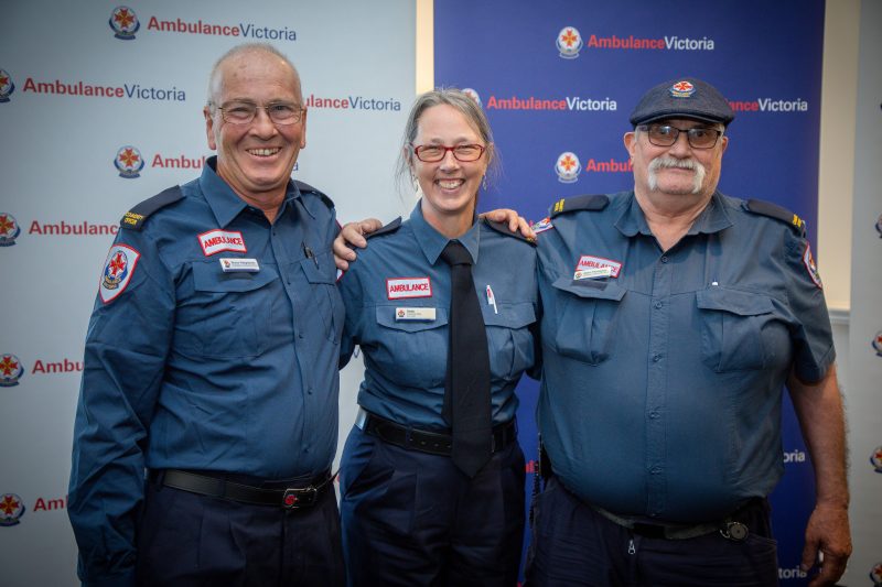 Three people in Ambulance Victoria paramedic uniform smile for the camera, in front of Ambulance Victoria banners.