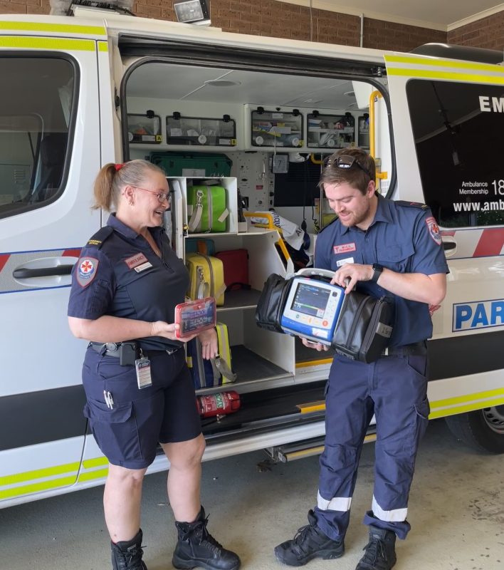 A woman holds a small CellAED in her hand. It's about the size of a small novel. A man holds a big, bulky defibrillator which requires two hands to lift up.