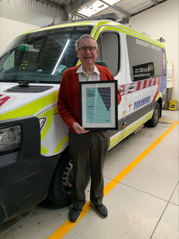 A man holds a framed certificate and smiles for the camera. He is standing in front of an ambulance.