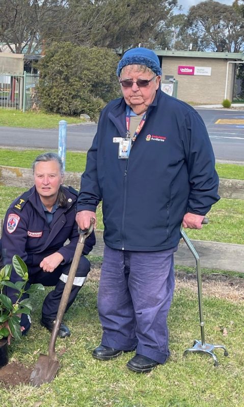 A woman crouches next to a small tree. A man with a walking stick stands next to her, holding a shovel.