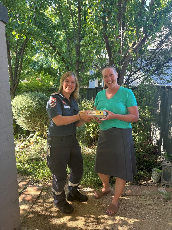 A woman paramedic and a woman in casual clothes hold a container of eggs and smile for the camera. They're in a garden, with trees behind them.