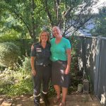 A woman paramedic and a woman in casual clothes smile for the camera, with their arms around each other. They're in a garden, with trees behind them.