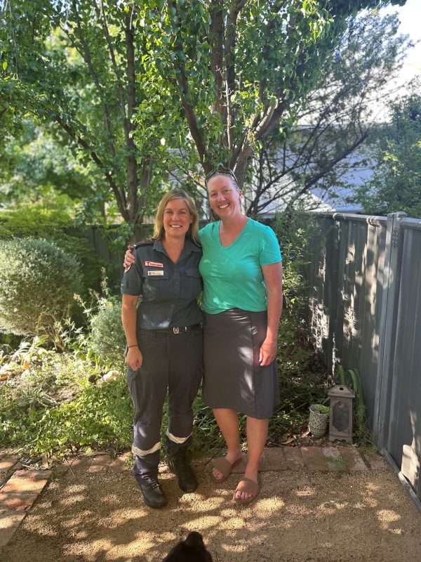 A woman paramedic and a woman in casual clothes smile for the camera, with their arms around each other. They're in a garden, with trees behind them.