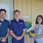 A paramedic and firefighter stand beside a woman in casual clothing, who holds an AED.