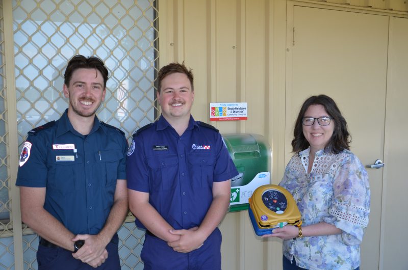 A paramedic and firefighter stand beside a woman in casual clothing, who holds an AED.