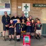 Four adults and seven children smile for the camera, with an AED and AED cabinet. They are at the 1st Kyabram Scout Group Hall.