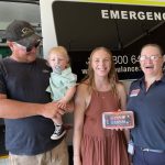 A man holding a toddler, a woman in a dress and a woman in Ambulance Victoria uniform smile for the camera. They hold a small defibrillator called a CellAED.