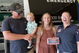 A man holding a toddler, a woman in a dress and a woman in Ambulance Victoria uniform smile for the camera. They hold a small defibrillator called a CellAED.