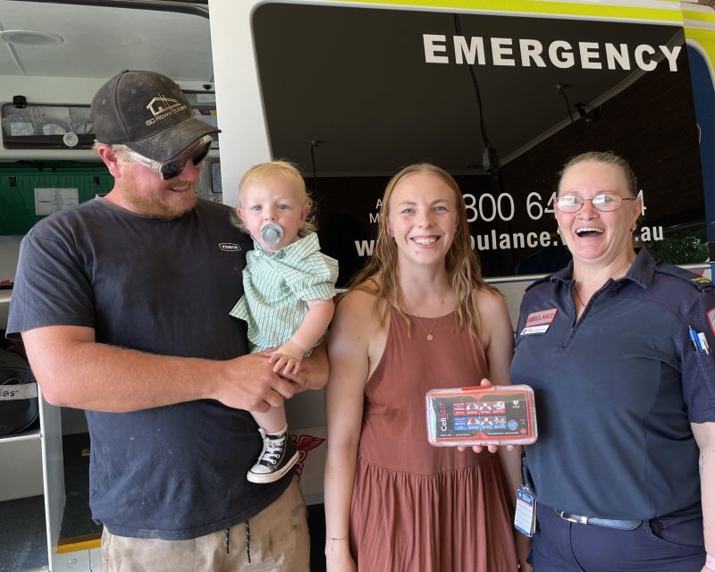 A man holding a toddler, a woman in a dress and a woman in Ambulance Victoria uniform smile for the camera. They hold a small defibrillator called a CellAED.