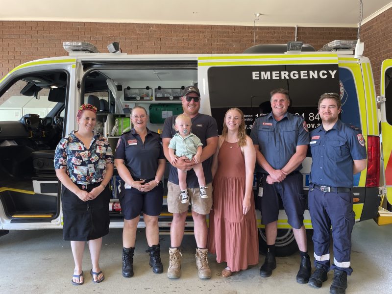 A group of people smile for the camera in front of an ambulance. Three wear Ambulance Victoria uniform. There is also a toddler being held.