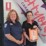 Two women standing in front of a sign. One is holding a defibrillator.