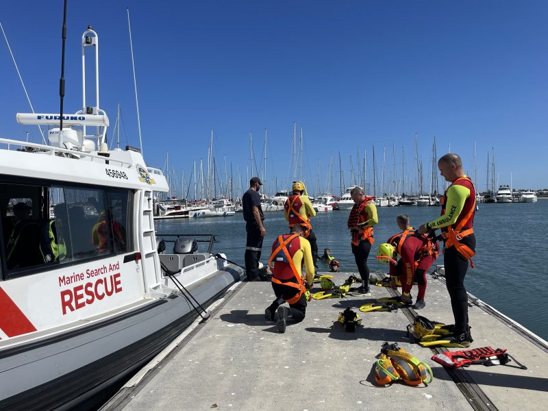 A group of paramedics in wetsuits standing beside a marine search and rescue boat preparing harnesses and life vests