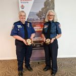 Marcia and Liz standing in uniform in front of an Ambulance Victoria banner holding awards