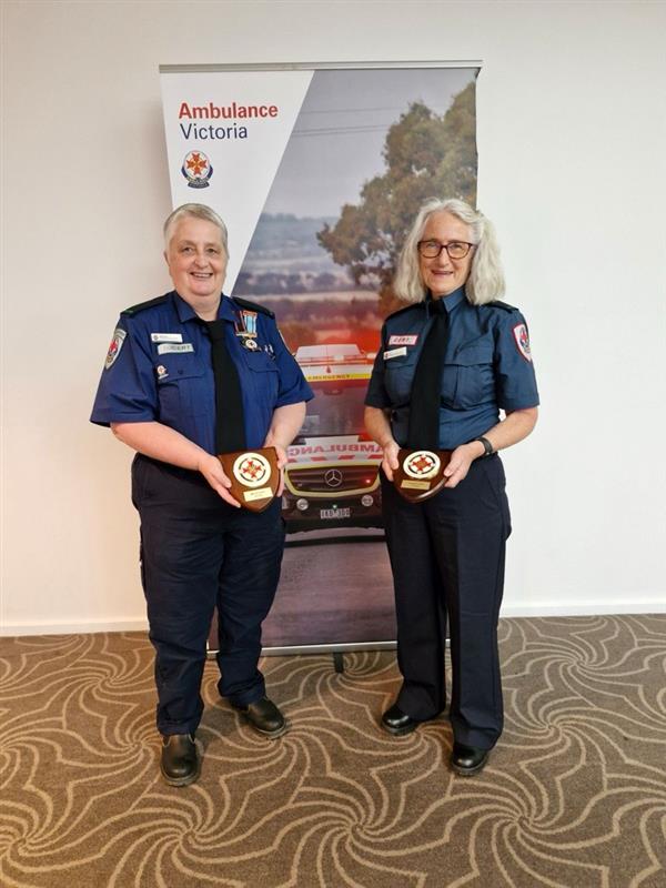 Marcia and Liz standing in uniform in front of an Ambulance Victoria banner holding awards