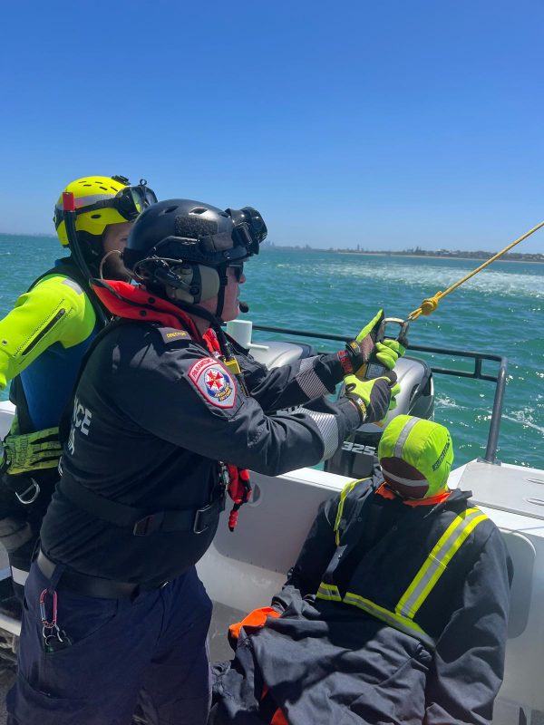 A paramedic in uniform on board a boat, holding the bottom of a winch rope to connect to a rescue mannequin