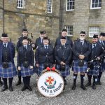 Members of a pipes and drums band standing in front of a brick building.