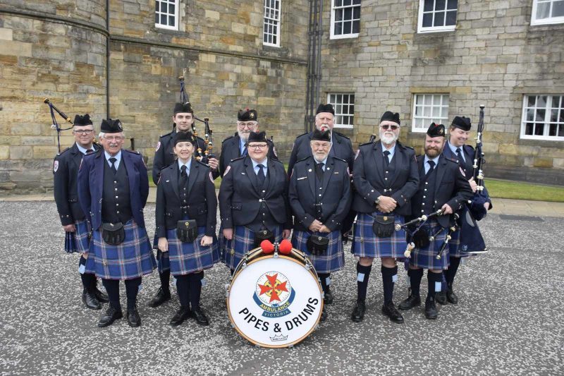 Members of a pipes and drums band standing in front of a brick building.