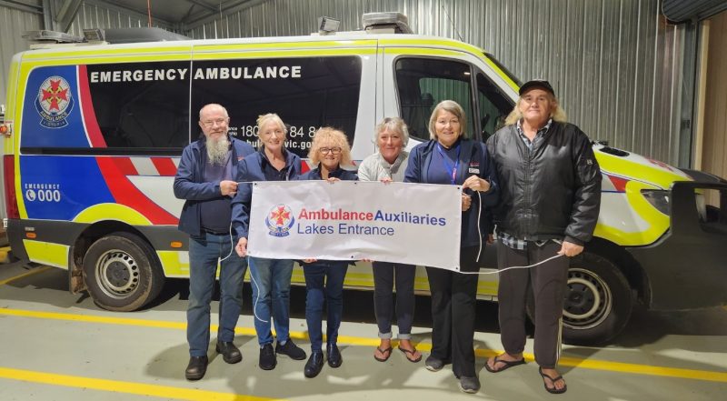 Six people stand in front of an ambulance, holding a banner which reads 'Ambulance Auxiliaries Lakes Entrance'.