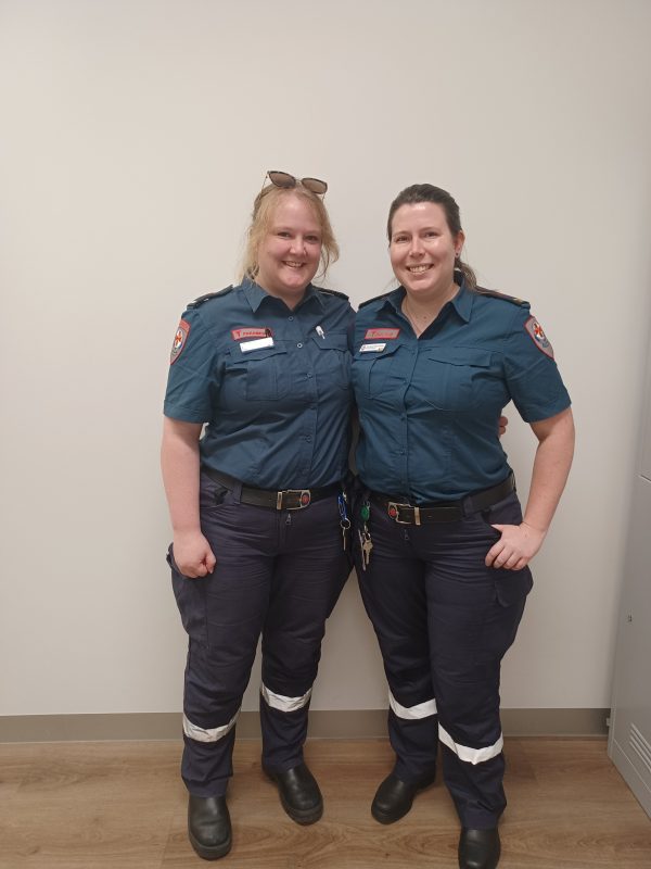 Two women in Ambulance Victoria uniform smile for the camera in front of a blank wall.