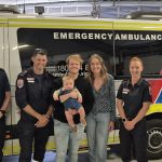 A group of people, including a baby, smiling in front of an ambulance.