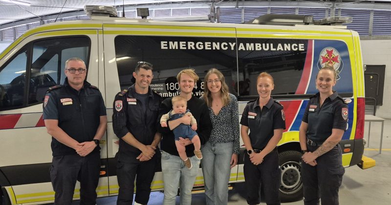 A group of people, including a baby, smiling in front of an ambulance.